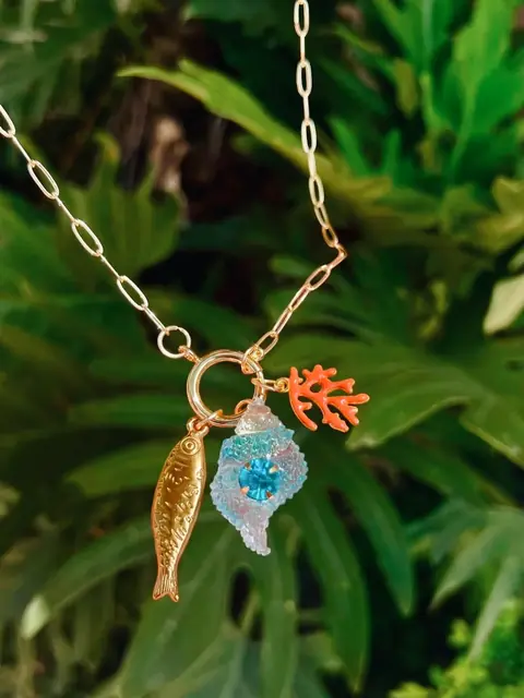 Cadena dorada con un pez dorado, un coral naranja y una caracola de vidrio azul, sobre hojas verdes